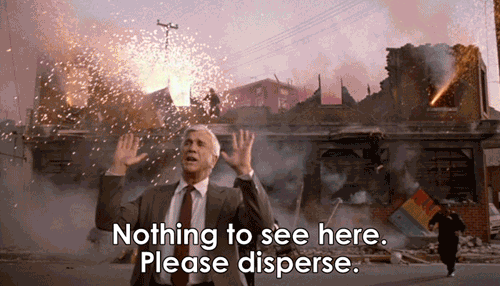 A white-haired white man in a suit gestures calmingly while in a bombed-out building behind him, fireworks go off and an explosion happens while people run away from it. He says "Nothing to see here. Please disperse."