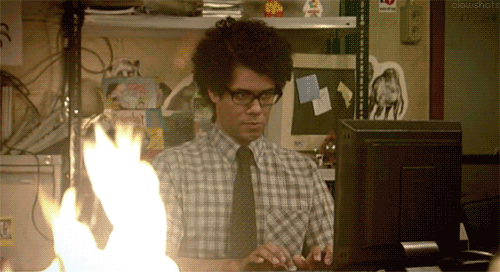 Moss from The IT Crowd — a black man with glasses and a tie on over a plaid shirt — looks at a monitor and types, and then looks at the fire burning in the foreground, then turns back to the monitor